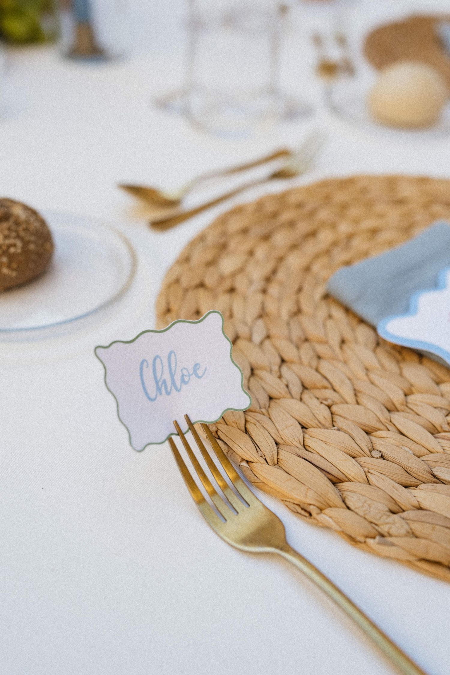 Gold fork and place card reading Chloe on woven rattan placemat at elegant wedding table setting.