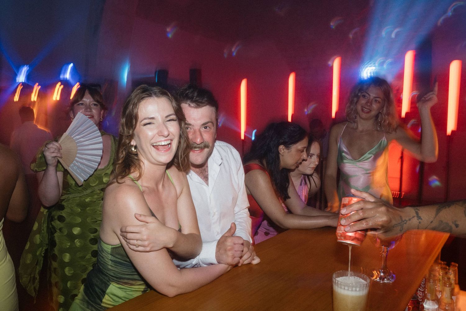 Couple smiling at a vibrant nightclub bar with colorful neon lights and party guests in the background.