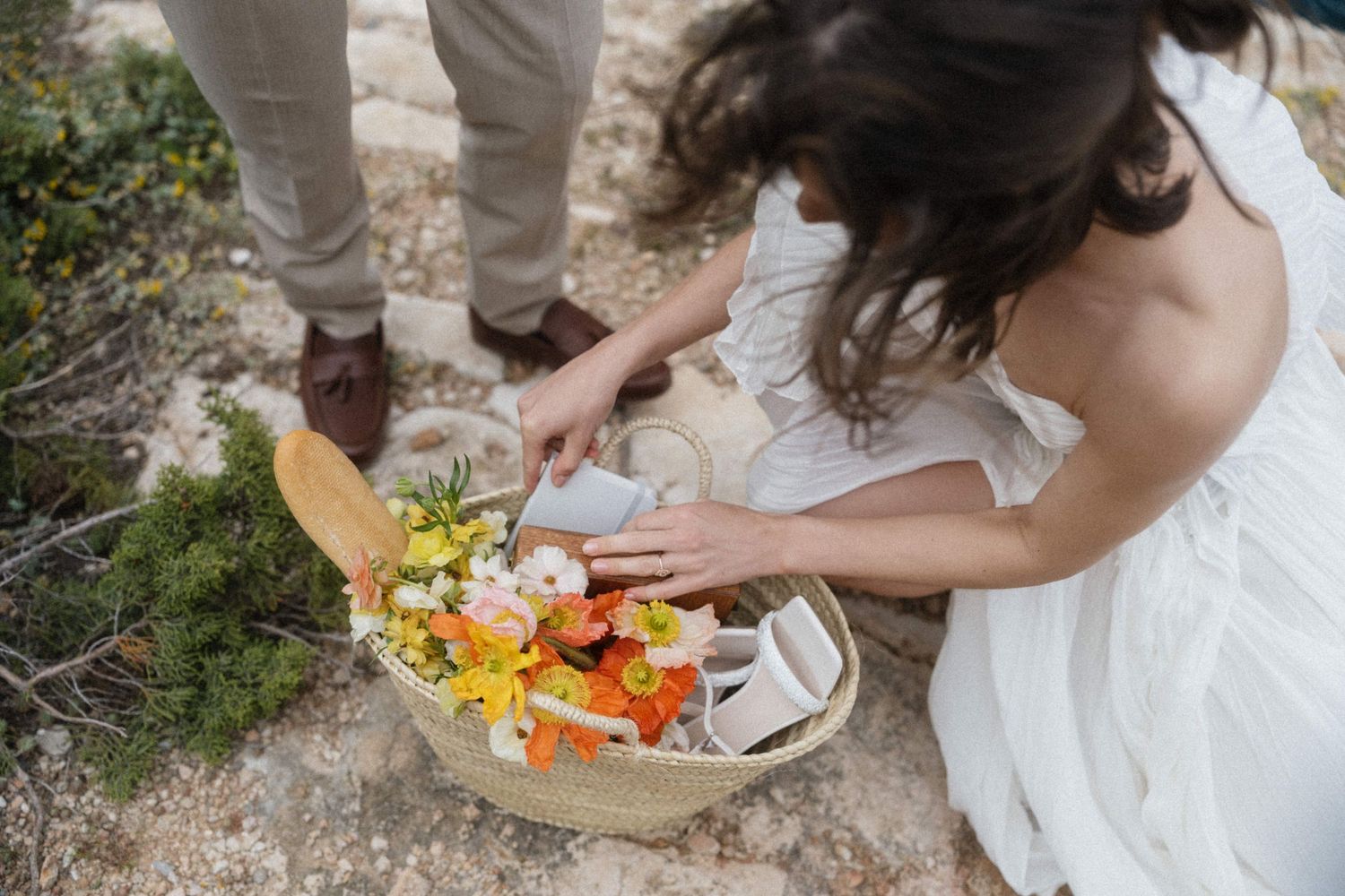 Bride in white dress arranging colorful flowers in a basket outdoors at a rustic wedding celebration.