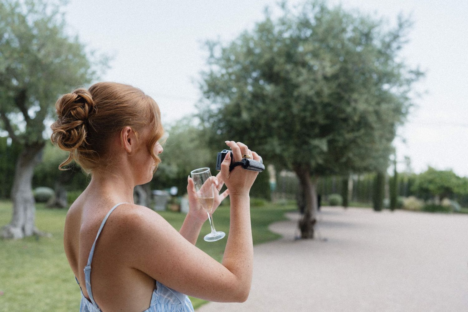Woman in blue dress holding champagne glasses outdoors in a park setting with lush green trees.
