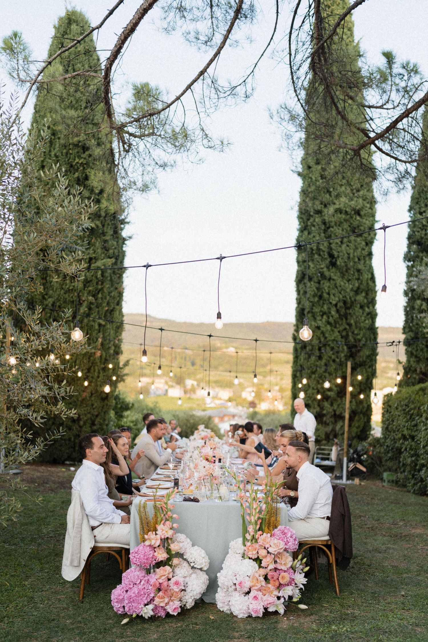 Elegant outdoor wedding reception with long floral table, string lights, and cypress trees in a garden setting.