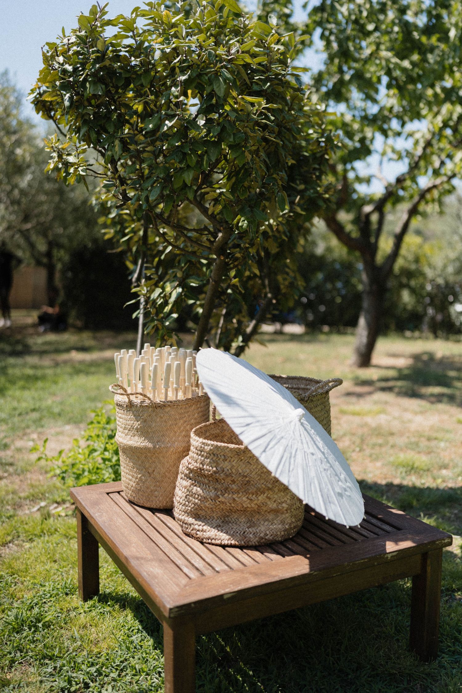 Wicker baskets and white parasol on wooden bench in sunny garden with lush green trees.