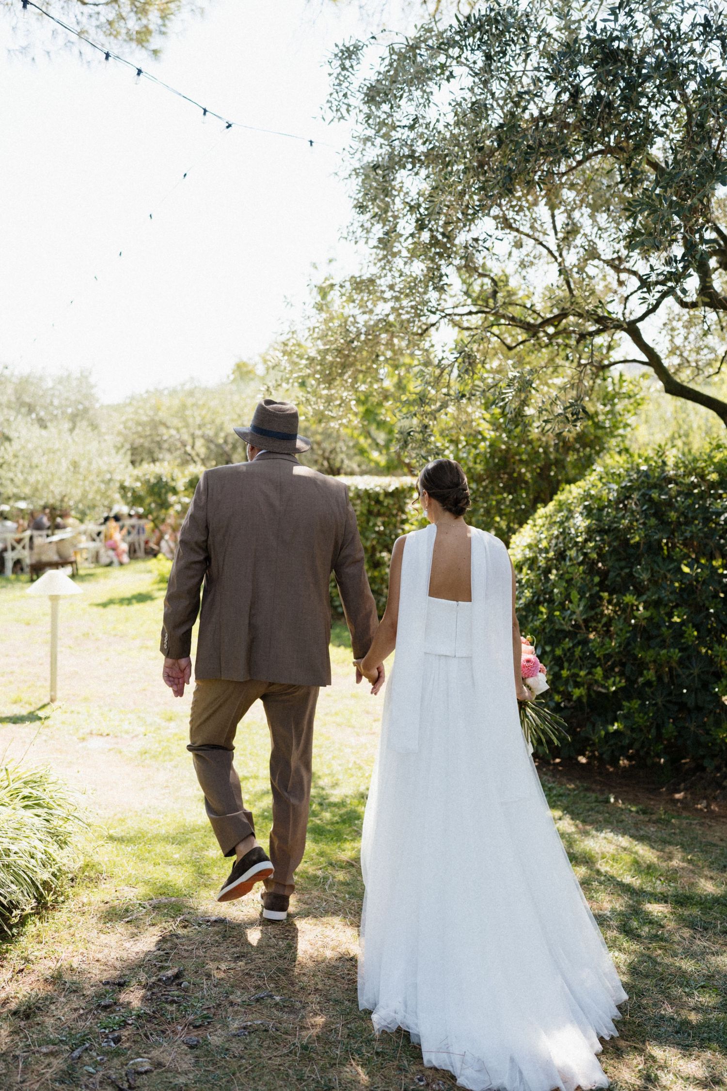 Bride in white gown and groom in brown suit holding hands walking through lush garden at outdoor wedding.