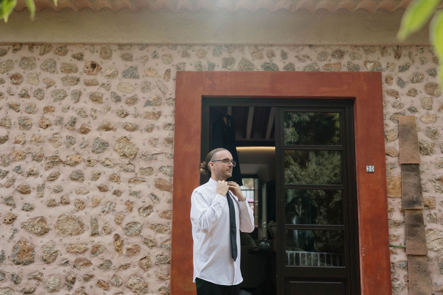 Man in a white shirt and dark tie adjusts his collar while standing in a red-framed doorway of a rustic stone building.