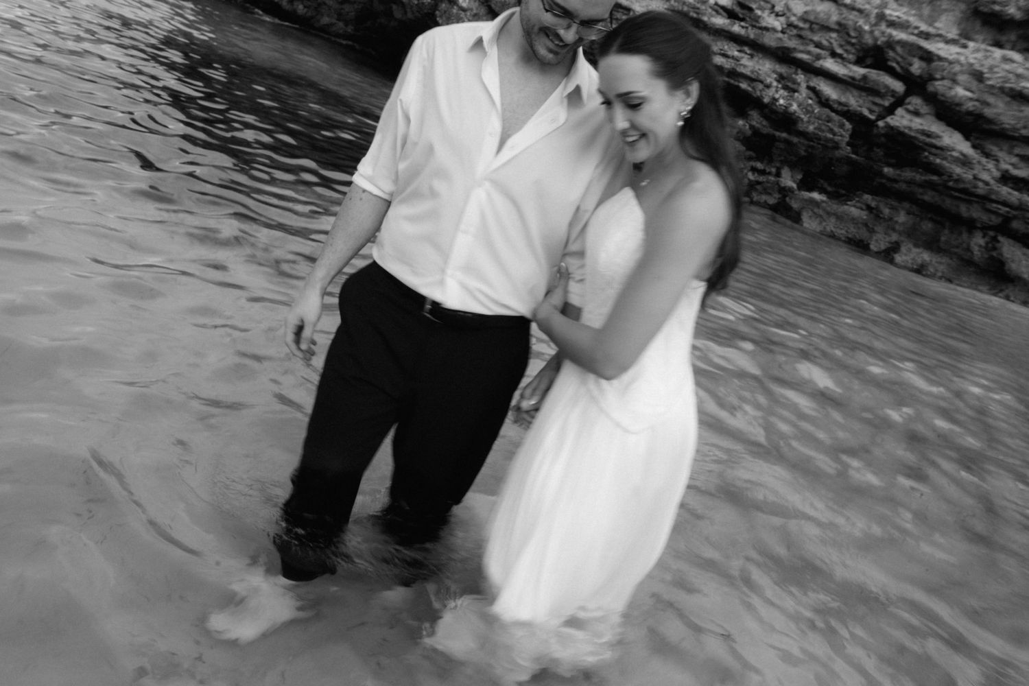 Black and white photo of a smiling couple embracing while standing in shallow ocean water near rocky cliffs, the bride in a white dress.