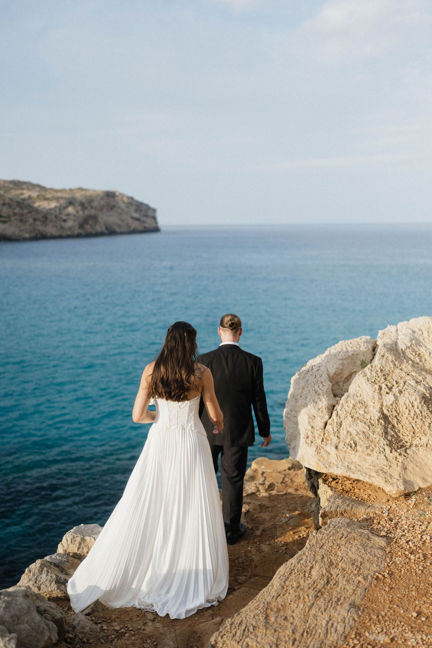 Bride in a flowing white dress and groom in a dark suit stand on a cliff edge gazing at the turquoise Mediterranean sea.