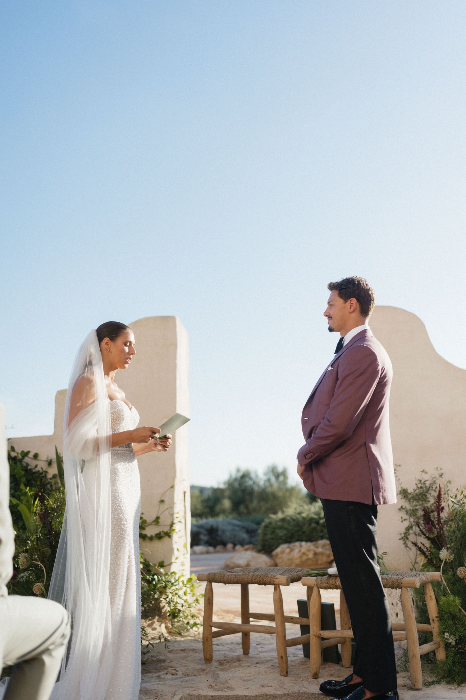 Bride reading her vows to the groom in a burgundy blazer during a sunny outdoor ceremony between whitewashed walls.