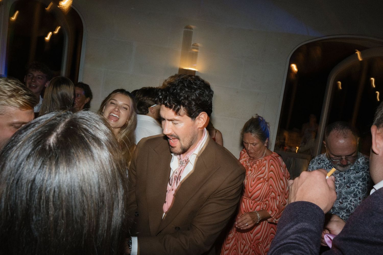 A man in a brown jacket and pink tie dances among laughing wedding guests in a warmly lit reception hall.