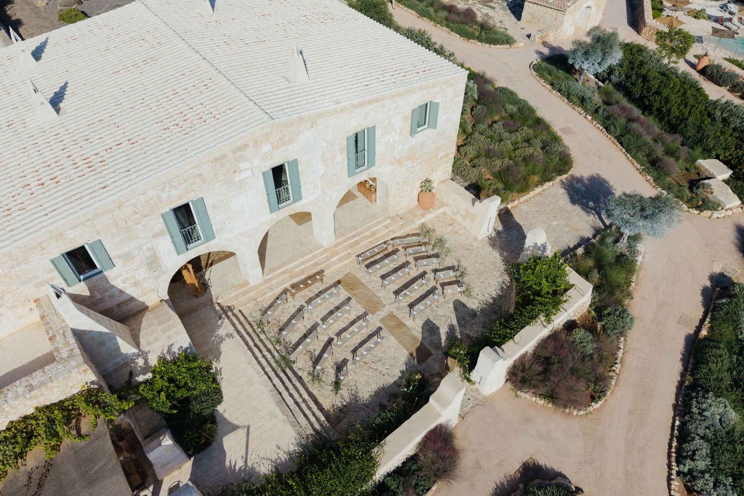 Aerial view of a whitewashed stone farmhouse with green shutters, arched loggia, wide stone steps and landscaped gardens.