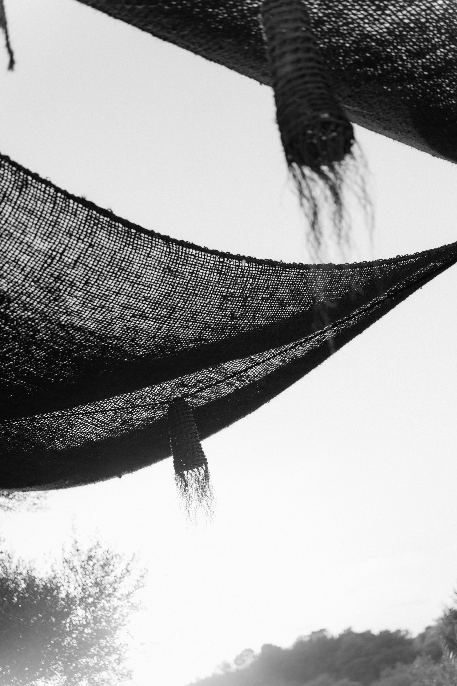 Black and white photo of woven shade sails with fringed tassels hanging against a bright sky, with silhouetted trees and hills below.