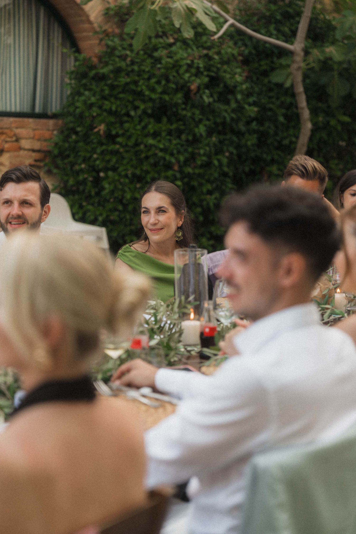 Wedding guests seated at decorated outdoor dinner table with candles and greenery.
