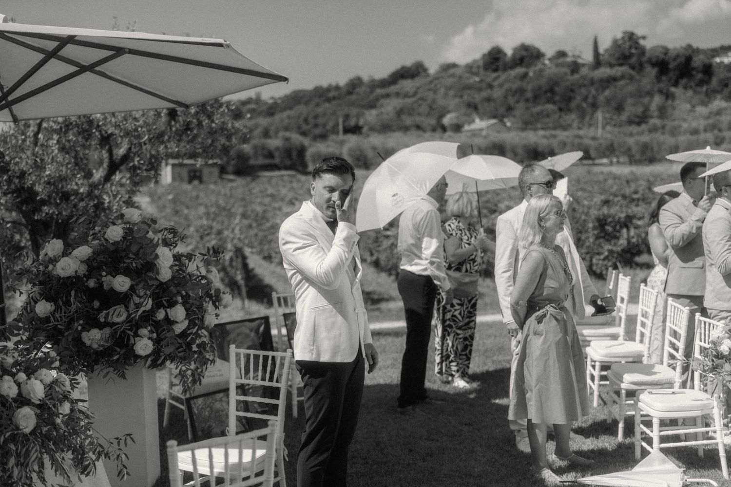 Elegant outdoor wedding ceremony with guests holding umbrellas amid vineyard backdrop in black and white.