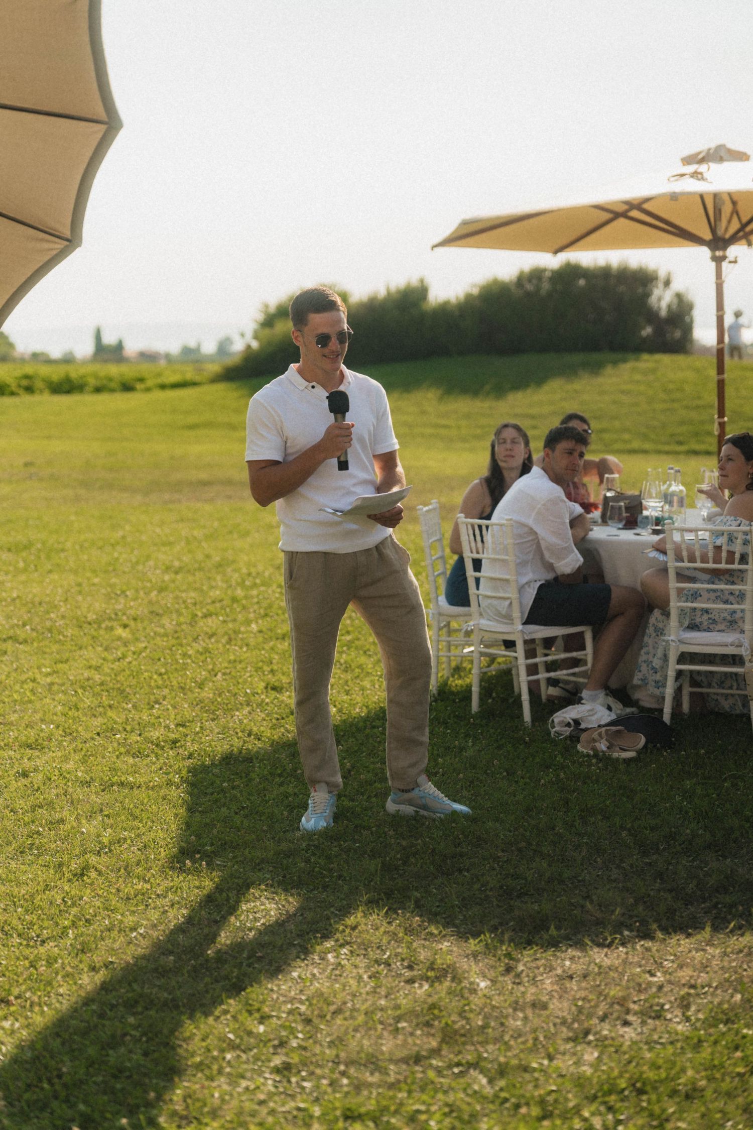 Man in white shirt gives a speech with a microphone at an outdoor wedding reception on a sunny green lawn.