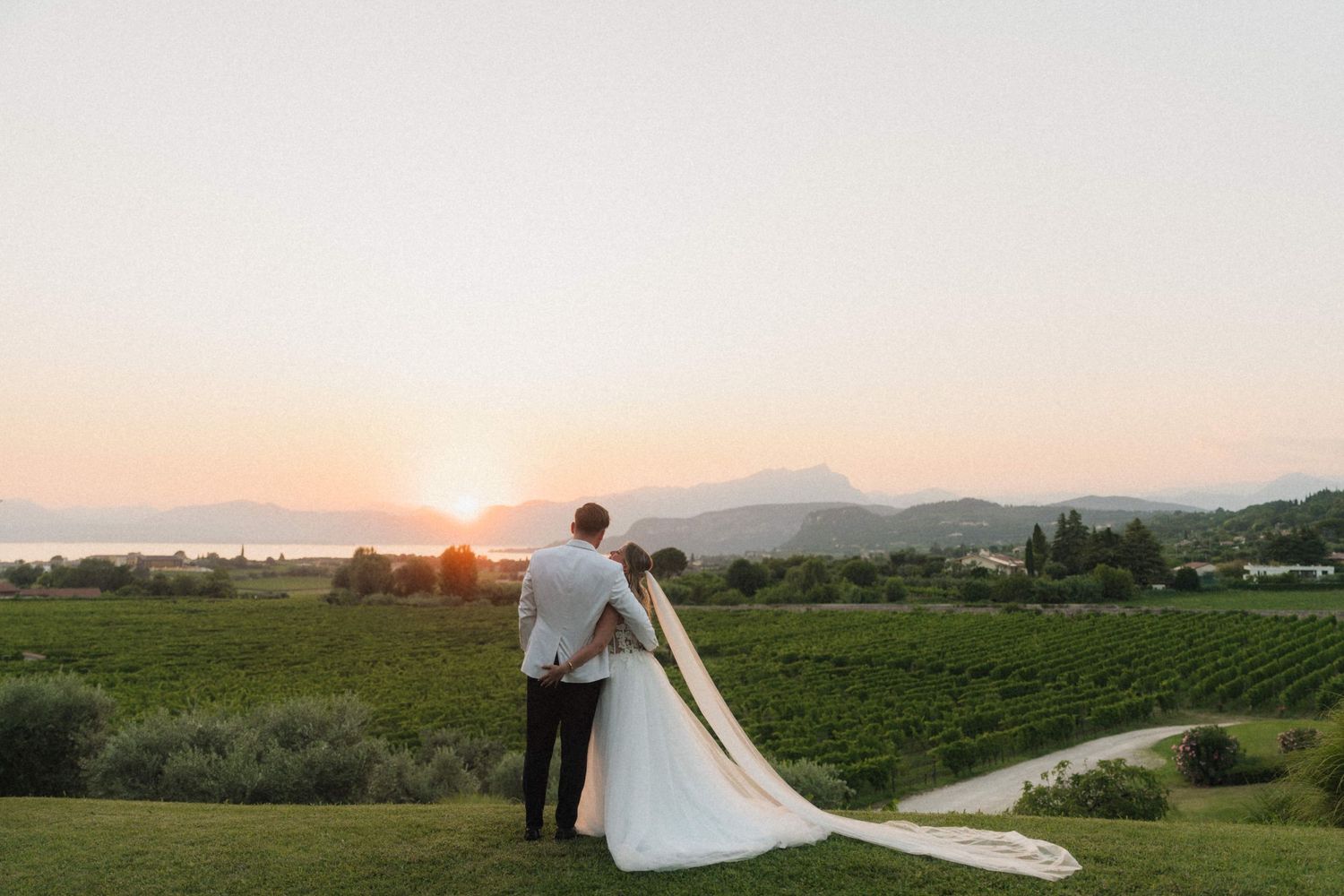 Bride and groom watching sunset over vineyard and mountains with long cathedral veil trailing.