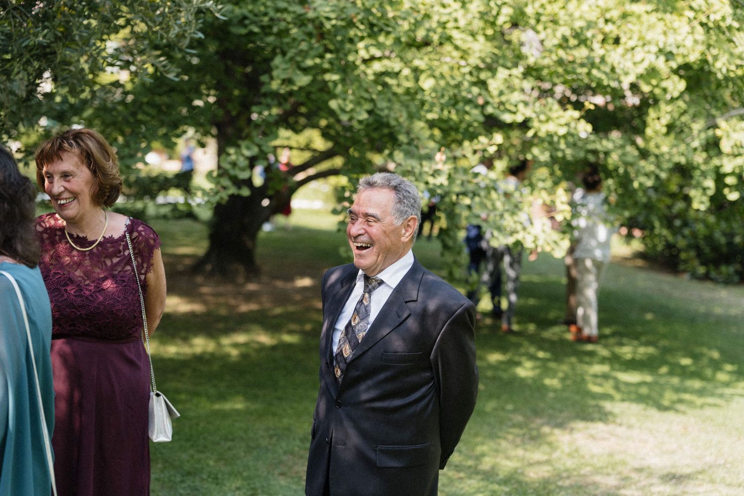 Elderly man laughing joyfully outdoors at a garden wedding celebration, wearing a dark suit with tie, surrounded by guests.