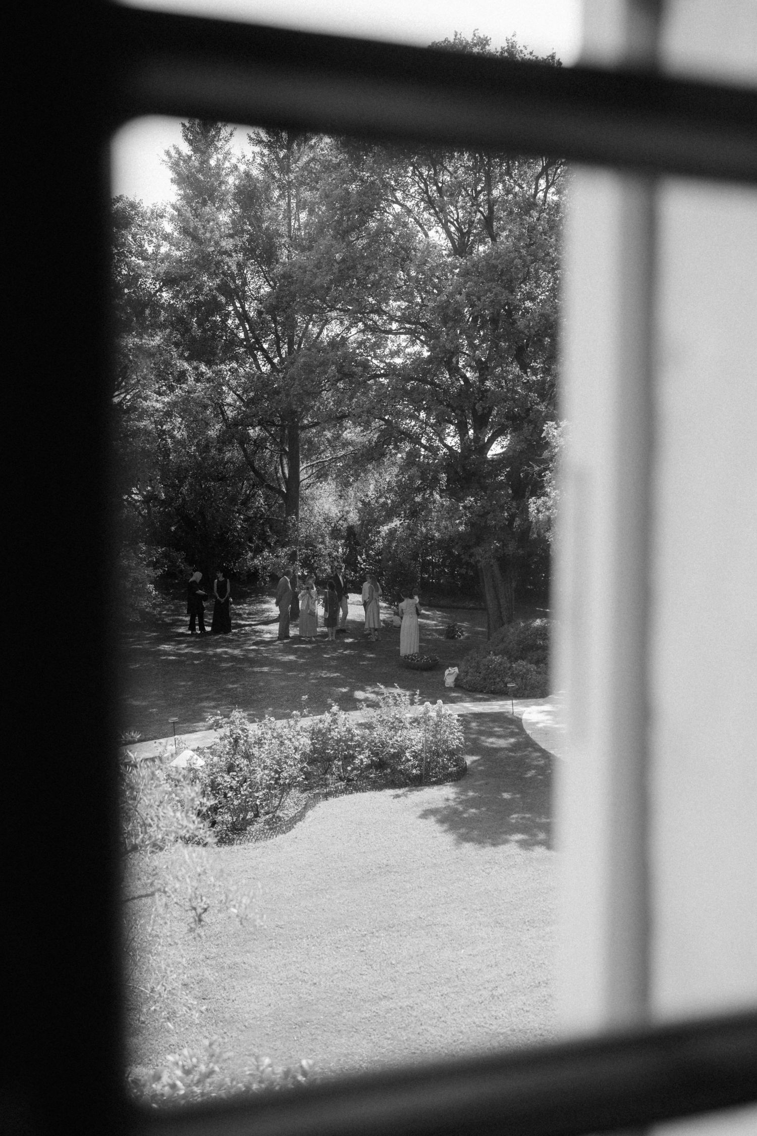Black and white view through window of wedding guests gathering in garden with trees.