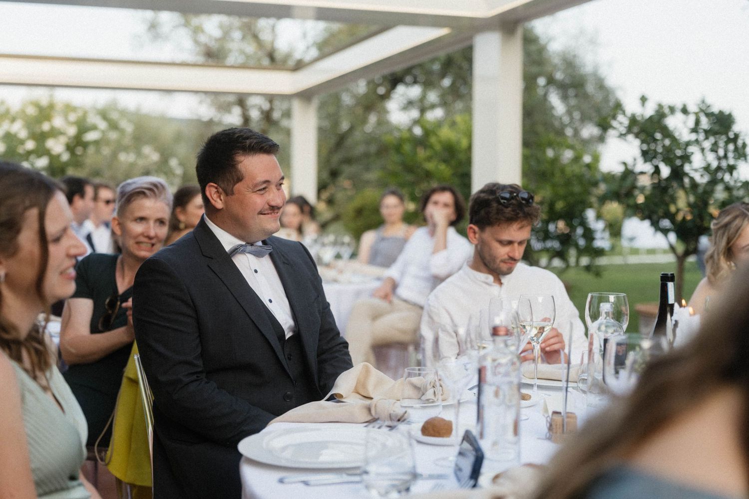 Elegant man in tuxedo smiling at wedding reception dinner table under outdoor pergola with guests.