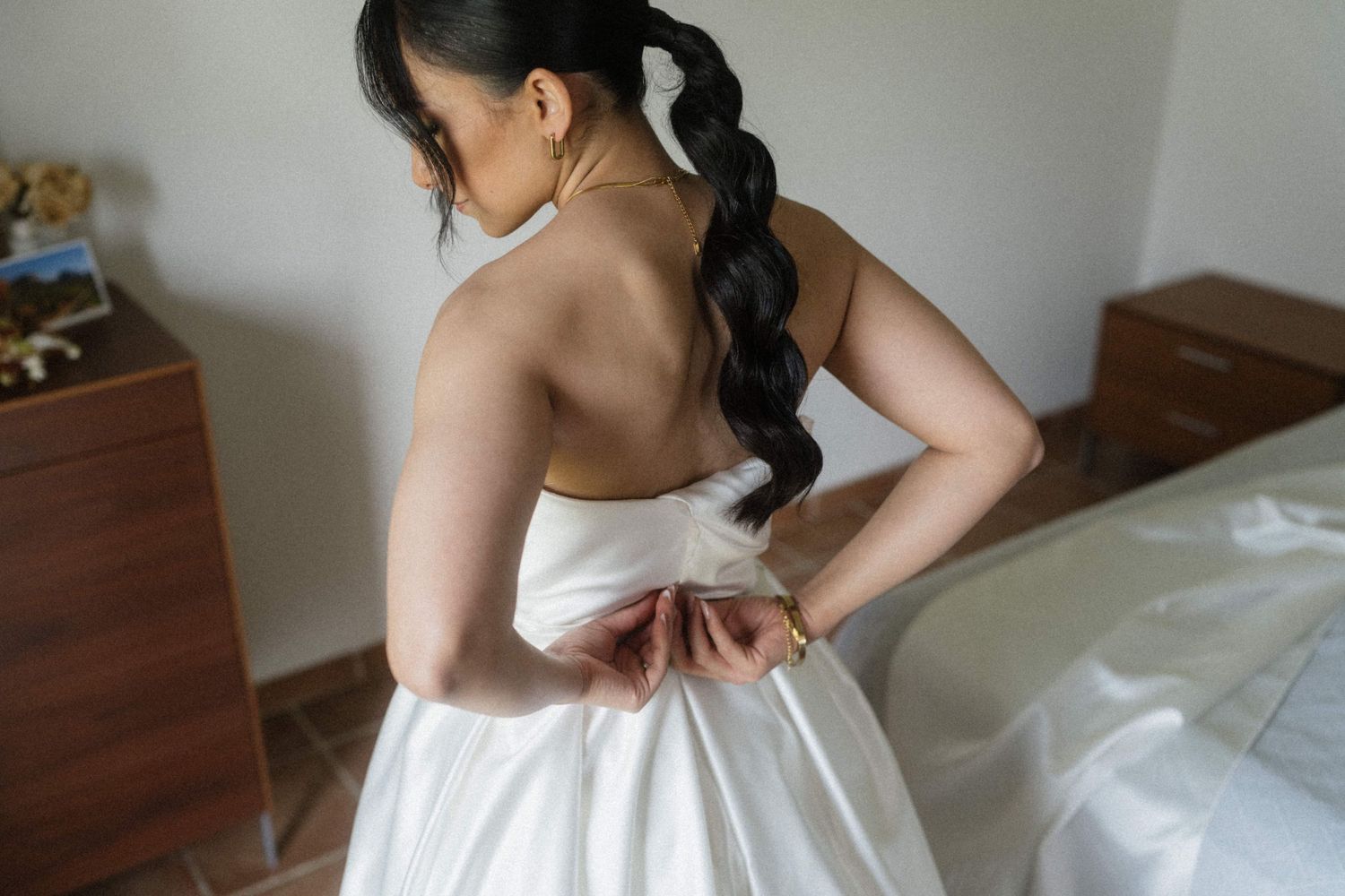 Bride in a white strapless wedding gown fastening the back of her dress in a sunlit bedroom.
