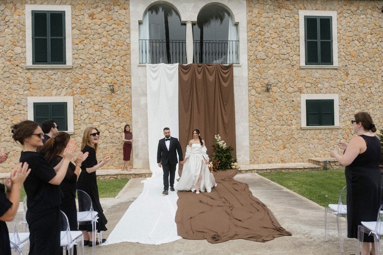 Newlyweds walk back down the aisle as guests applaud at elegant outdoor stone villa wedding.