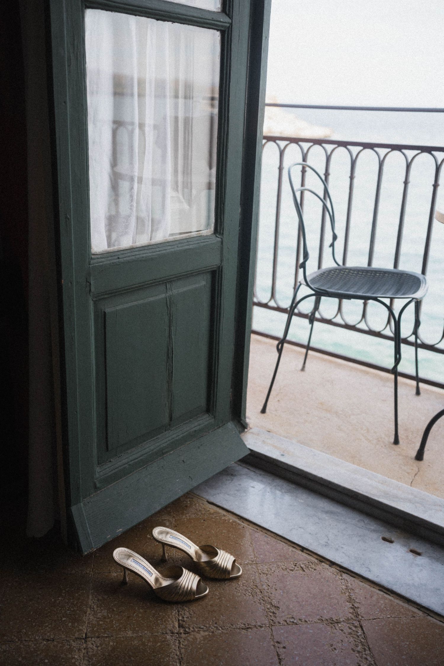Green vintage door open to iron balcony with bridal heels placed on tiled floor nearby.