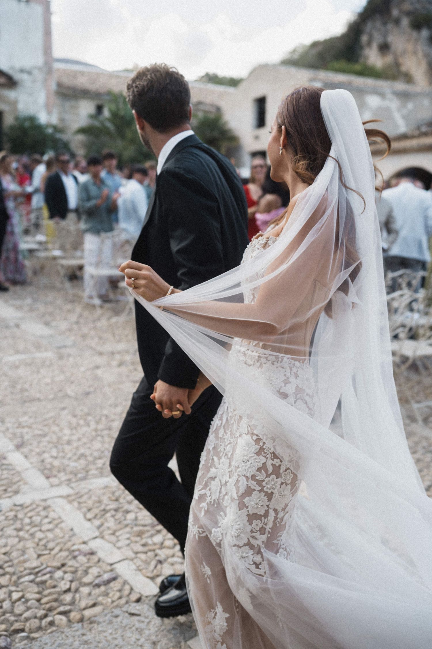 Bride in lace gown with flowing veil walks hand in hand with groom.