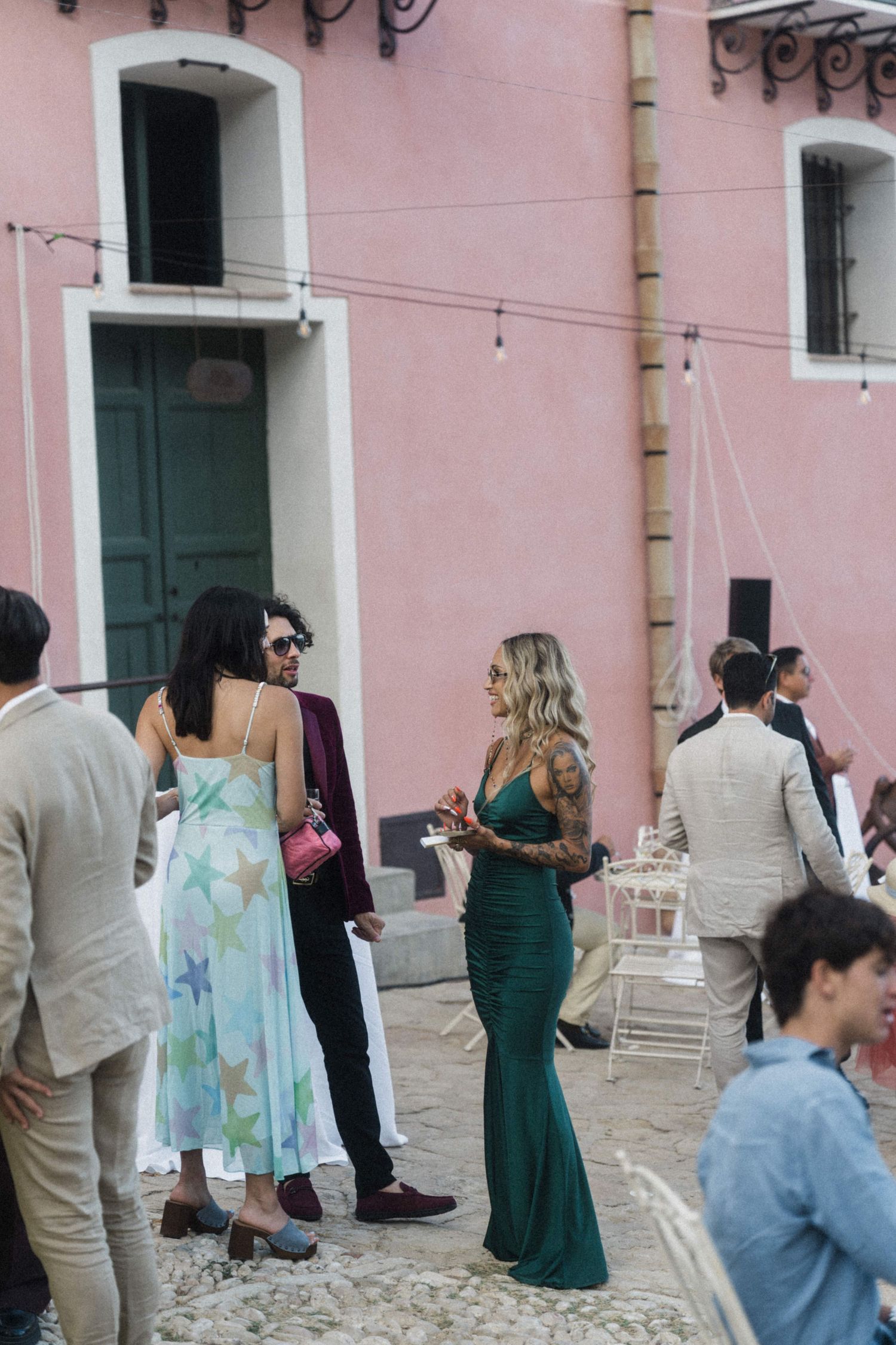 Wedding guests mingling outdoors near pink building, including woman in green gown and couple in colorful attire.