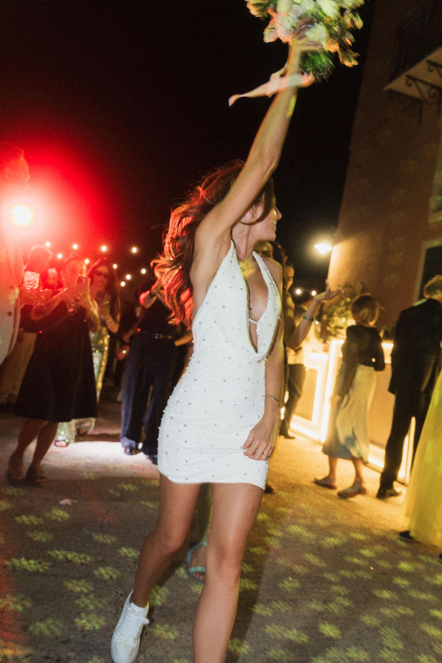Bride in white mini dress tossing bouquet into air at outdoor wedding reception at night.