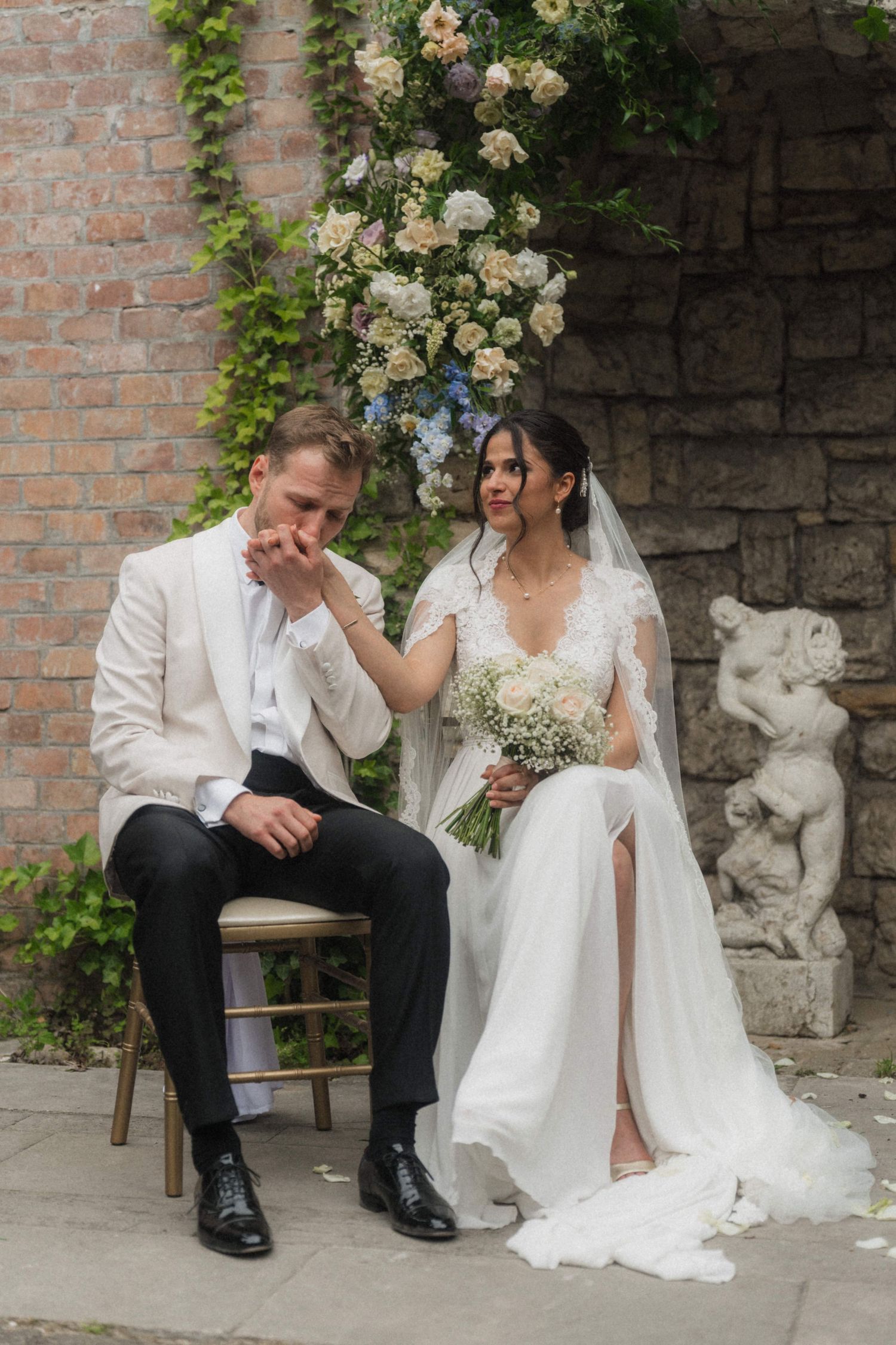 Bride and groom seated at outdoor wedding ceremony beneath floral arch against ivy-covered brick wall with cherub statue.