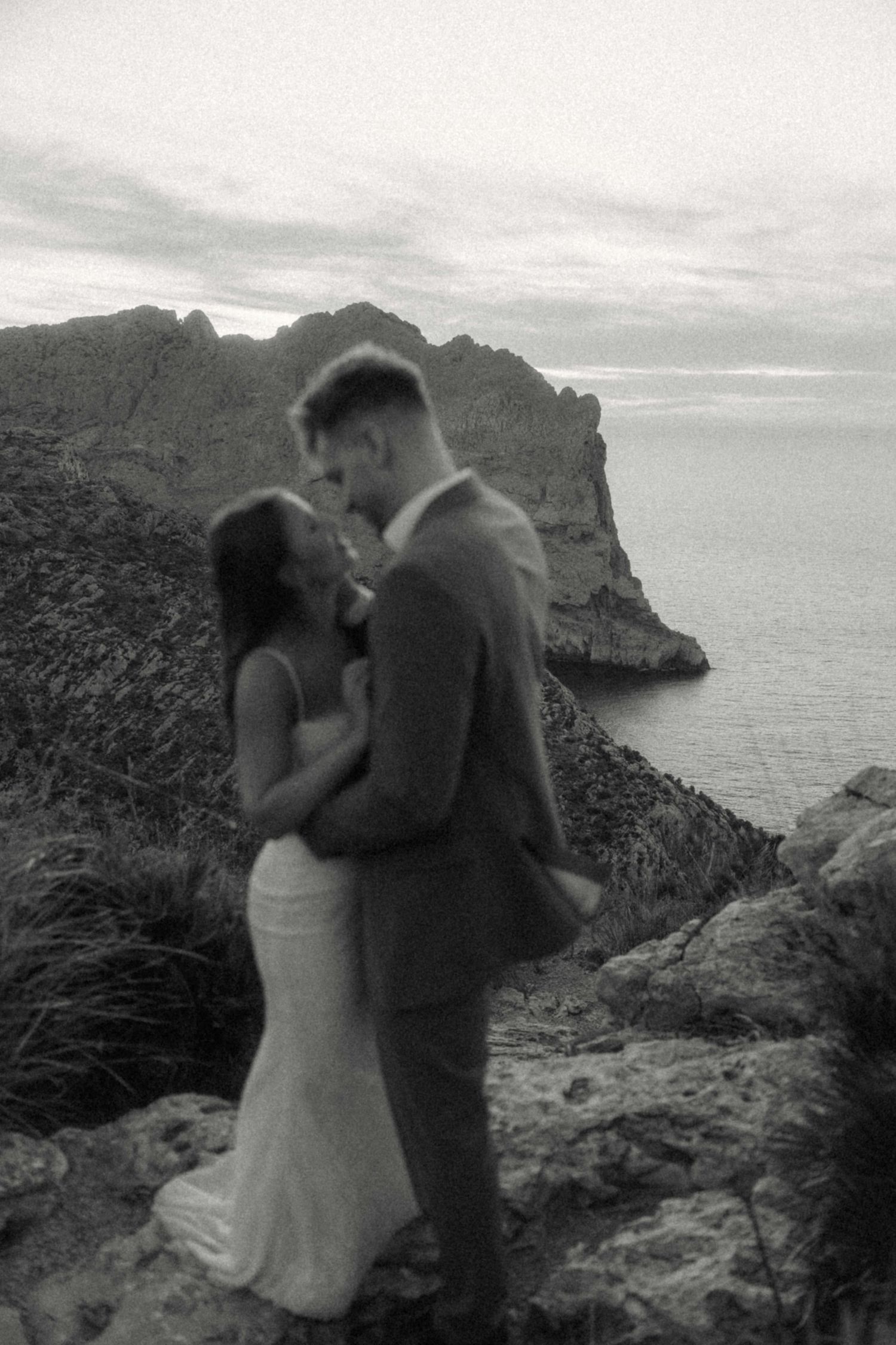Black and white photo of bride and groom embracing on dramatic coastal cliffs with mountain and sea views.