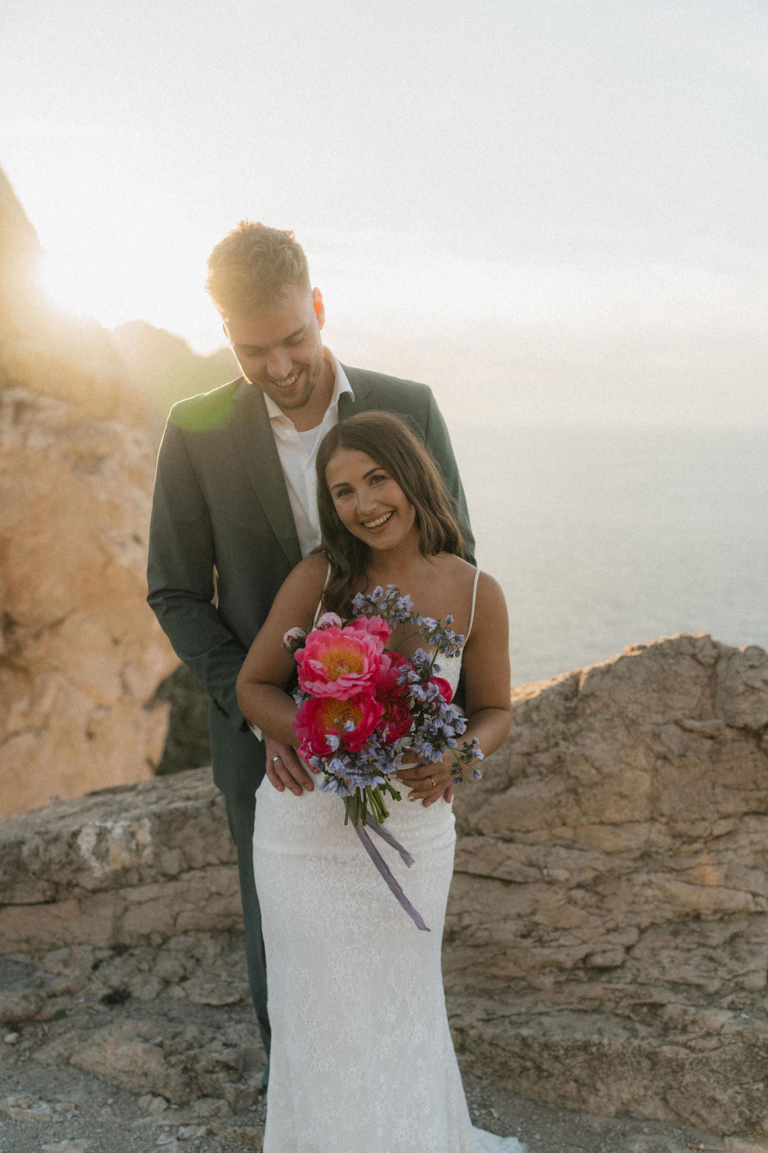 Groom embraces bride from behind at golden sunset overlooking the sea, bride smiling holding bright pink peony bouquet.