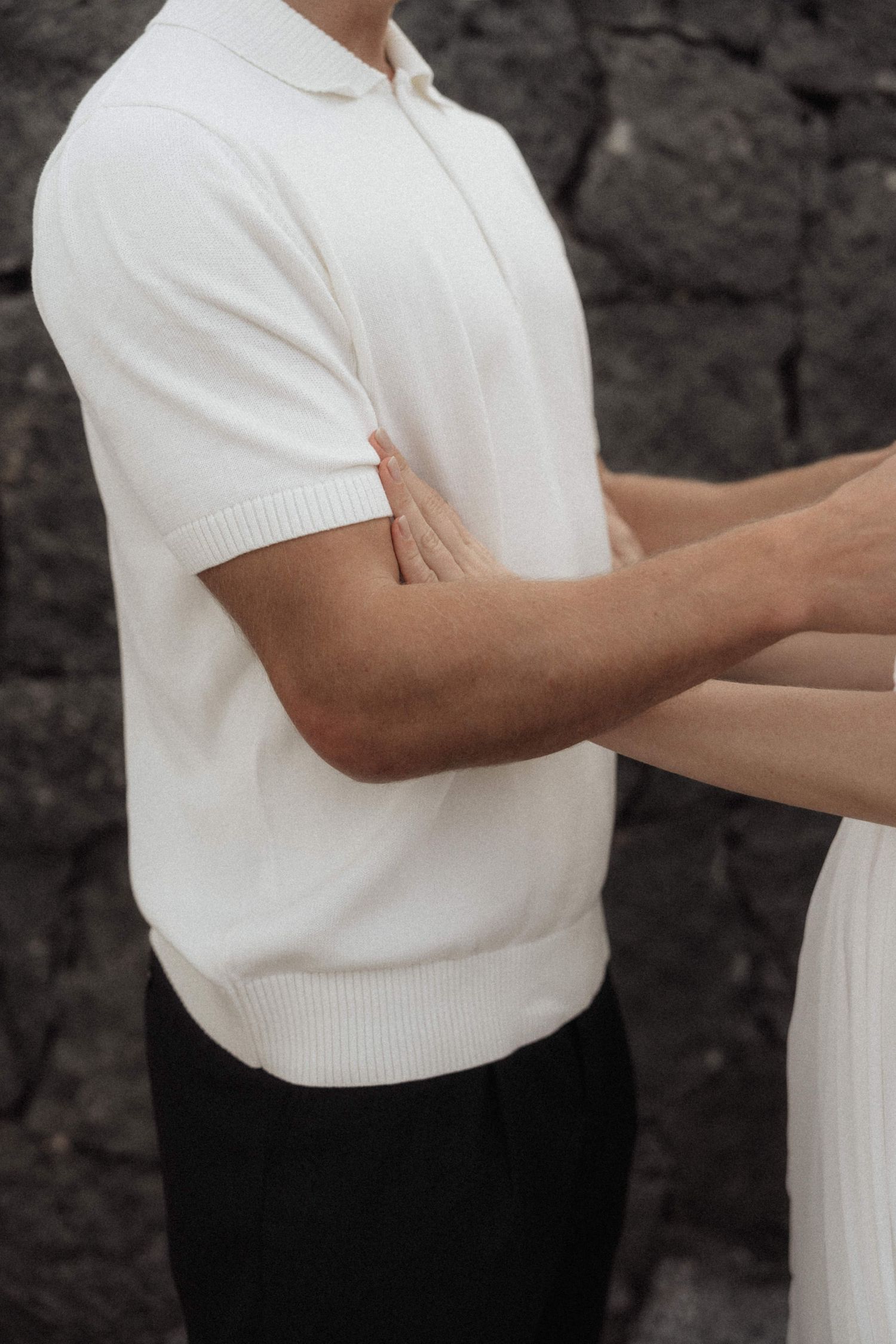 Groom in white knit polo shirt holds bride's hand against dark stone wall backdrop.