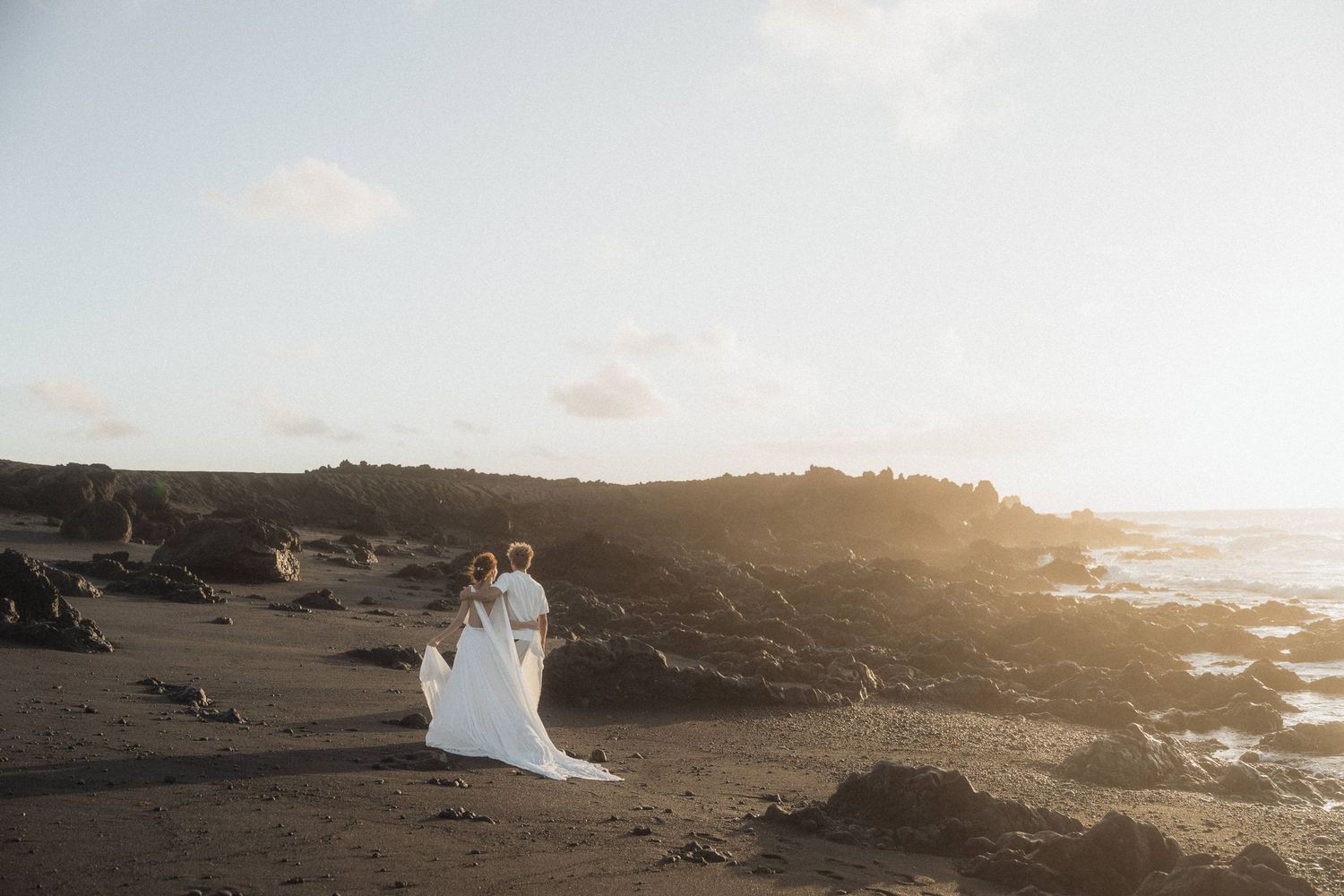 Wedding couple standing together on a volcanic black sand beach at golden hour sunset.