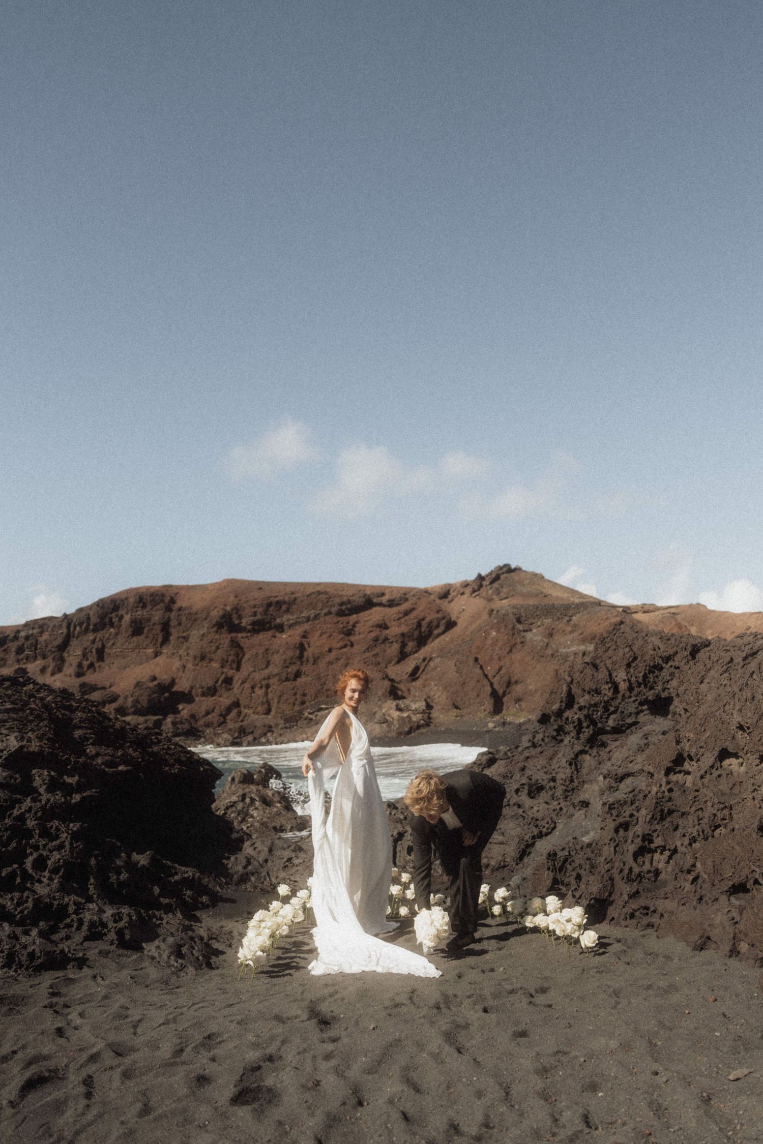 Bride in white gown stands on black volcanic beach with floral arch and rocky cliffs.