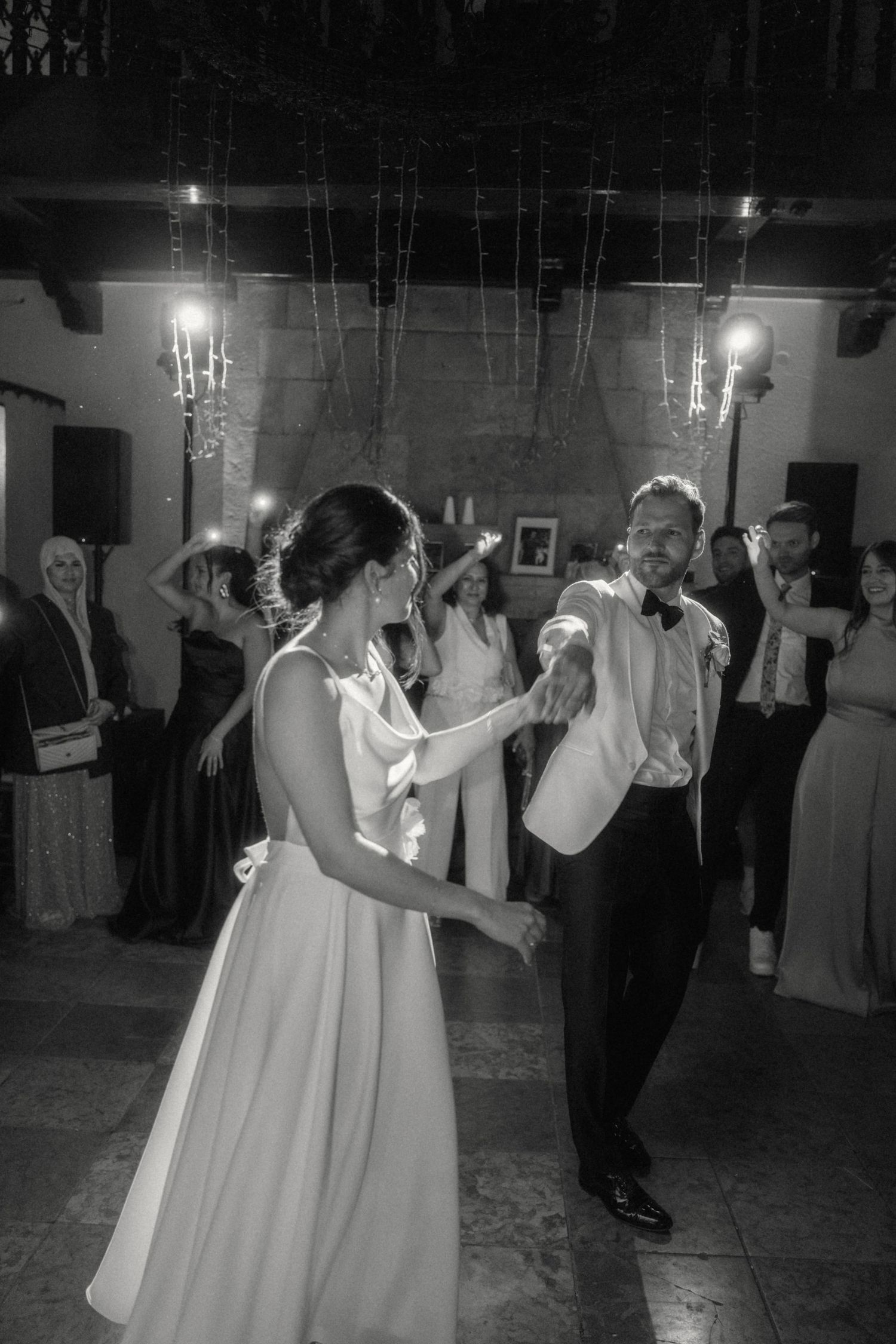 Bride and groom share first dance in elegant venue as guests celebrate in black and white photo.