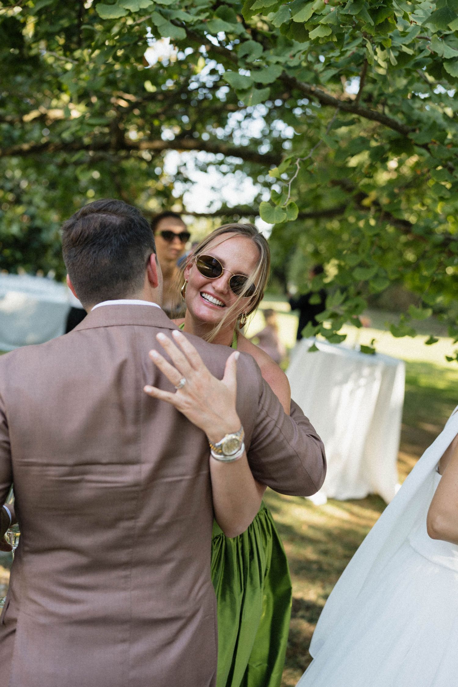 Woman in green dress and sunglasses smiling warmly at a wedding guest outdoors under green trees.