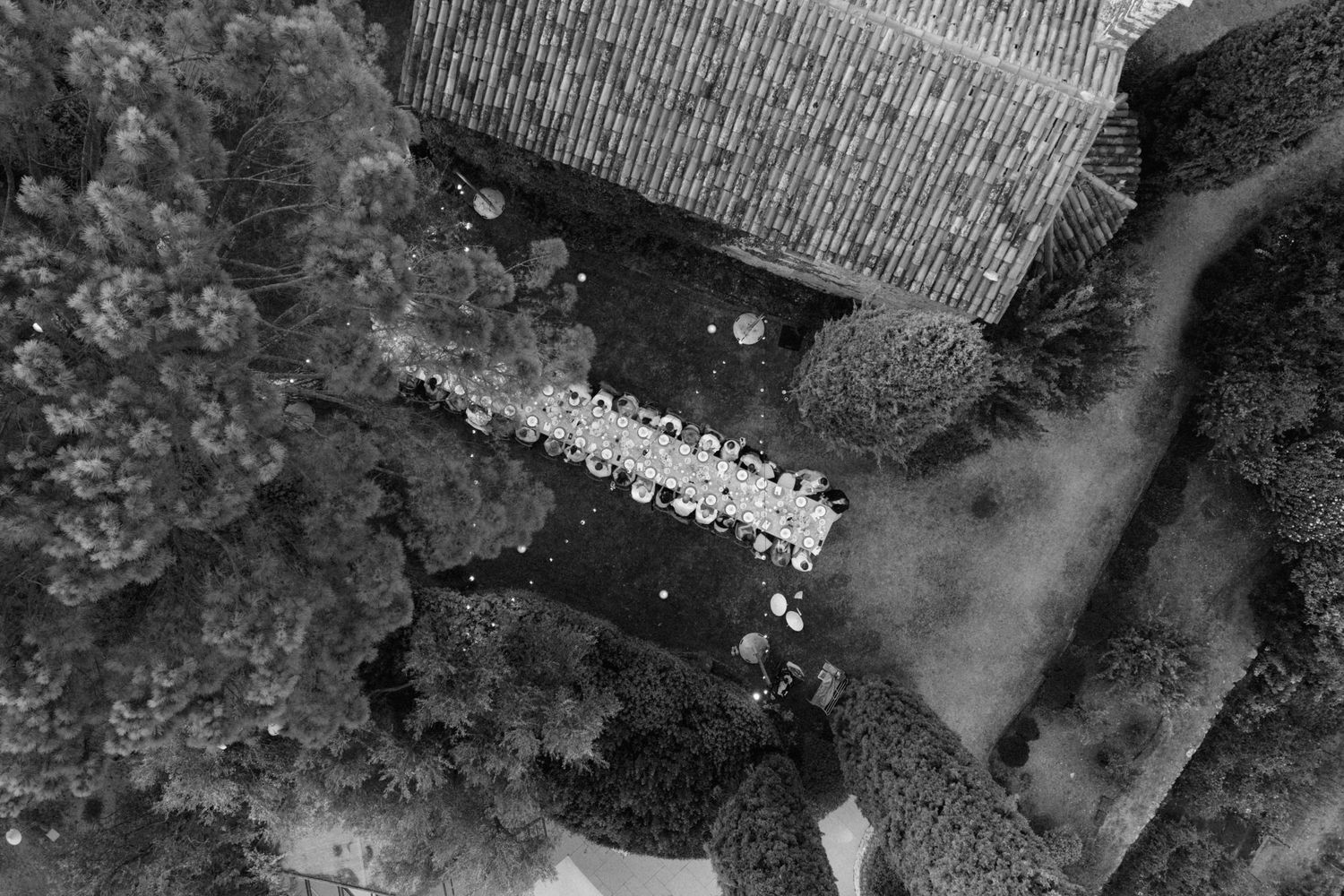 Black and white aerial drone view of a long outdoor wedding reception table beside a stone building.
