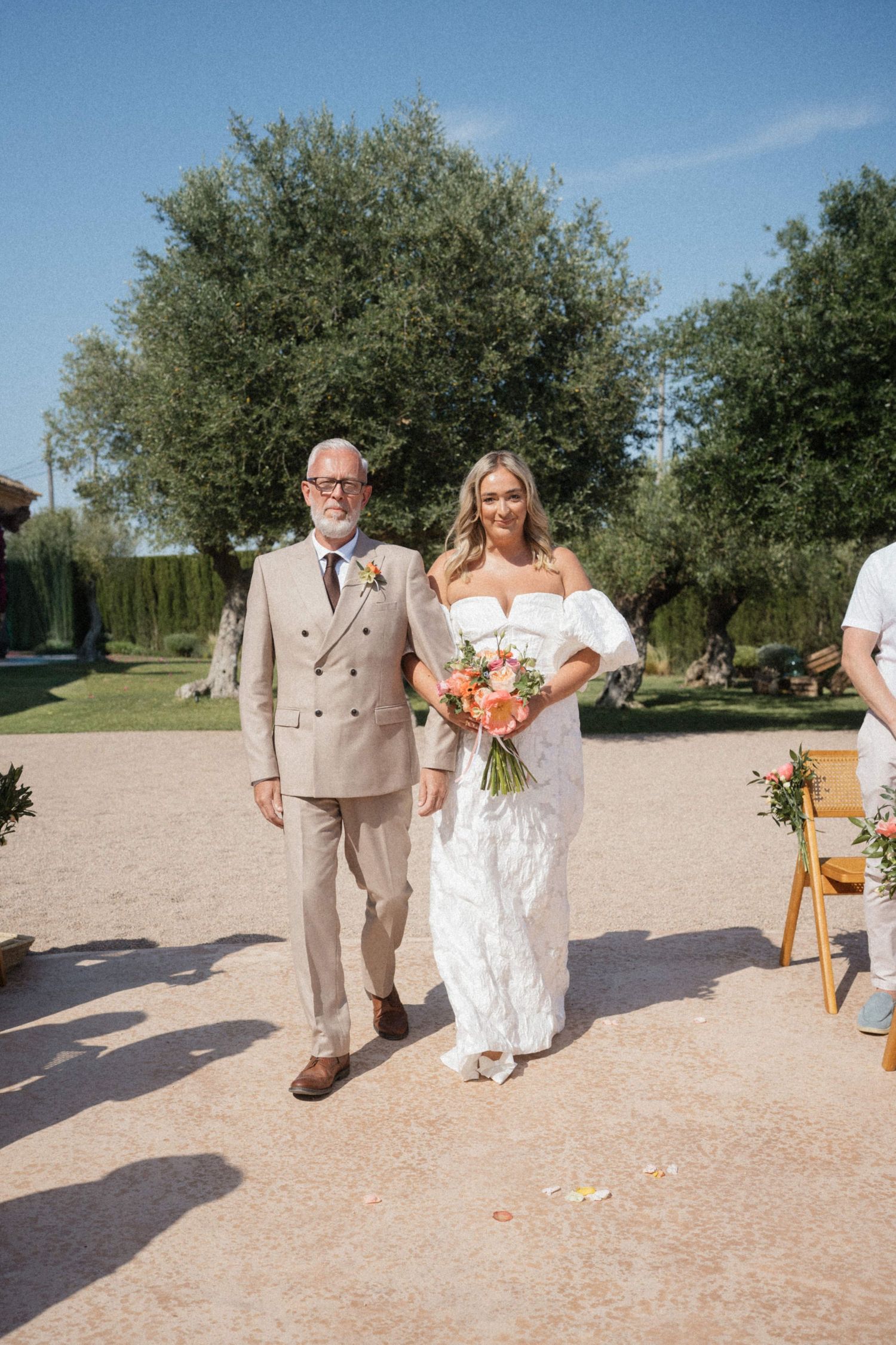 Bride in white off-shoulder gown walks down outdoor aisle with older man in beige suit holding orange bouquet.