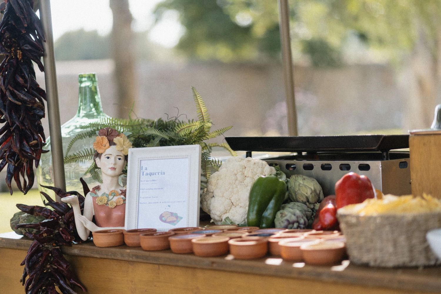 Rustic Mexican food display with clay bowls, fresh vegetables, chili pepper garlands, and decorative figurine on wooden shelf.