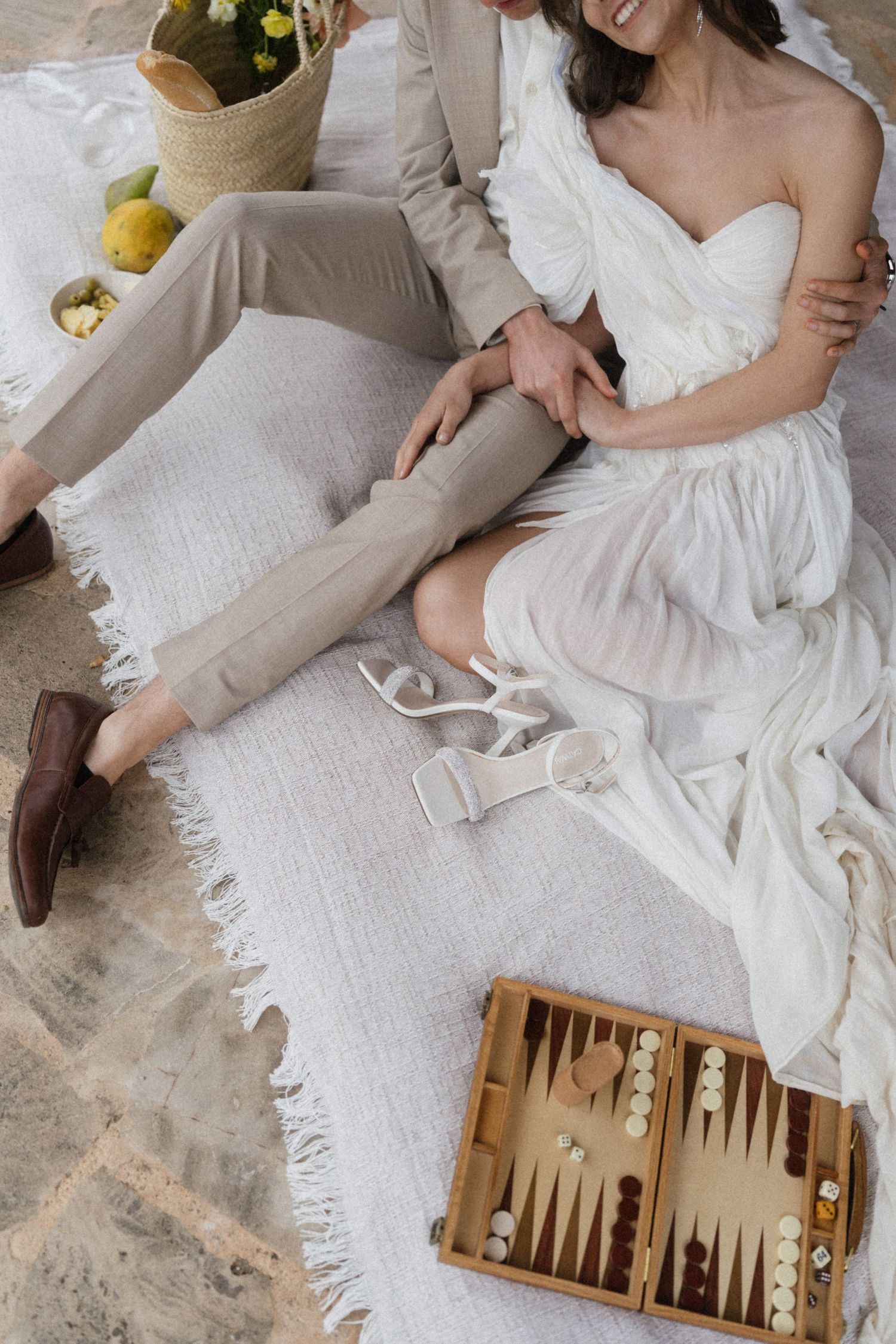Bride and groom sitting on white blanket with backgammon board during outdoor wedding picnic.