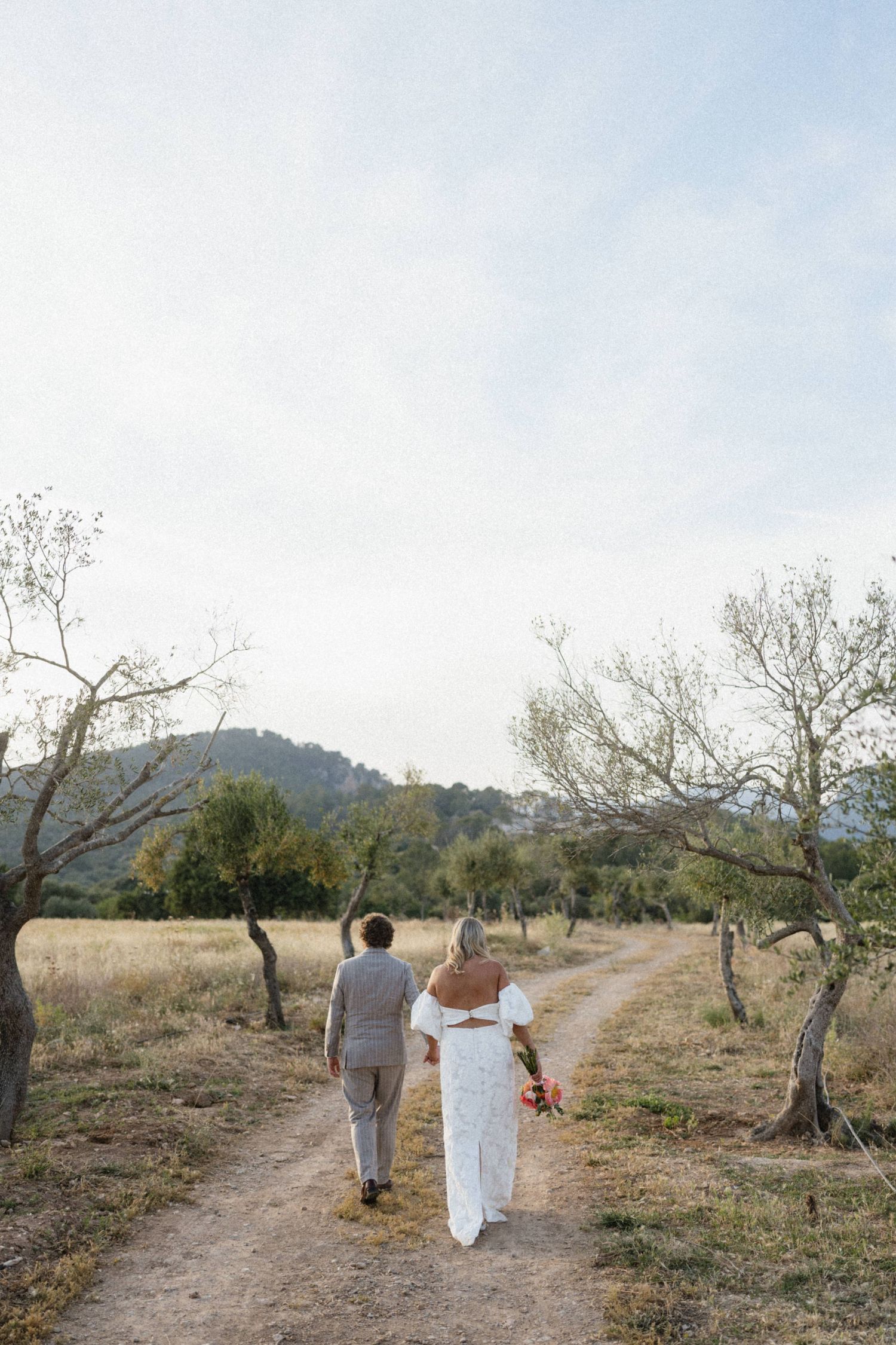 Bride and groom walking hand in hand down a dirt path through olive trees in a scenic landscape.