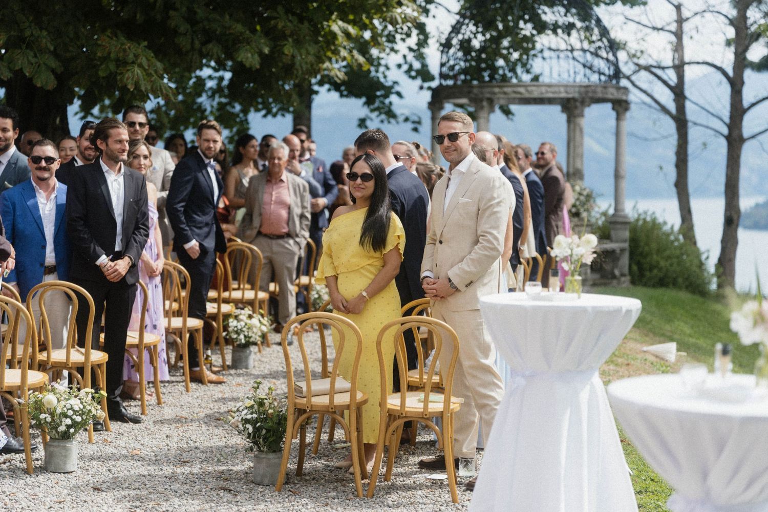 Wedding guests in formal attire seated in outdoor ceremony chairs, woman in yellow dress escorted down aisle.