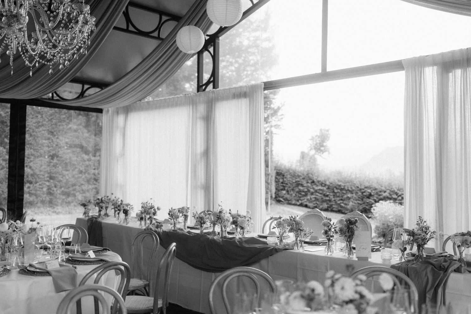 Black and white photo of elegant wedding reception hall with large windows and floral centerpieces.