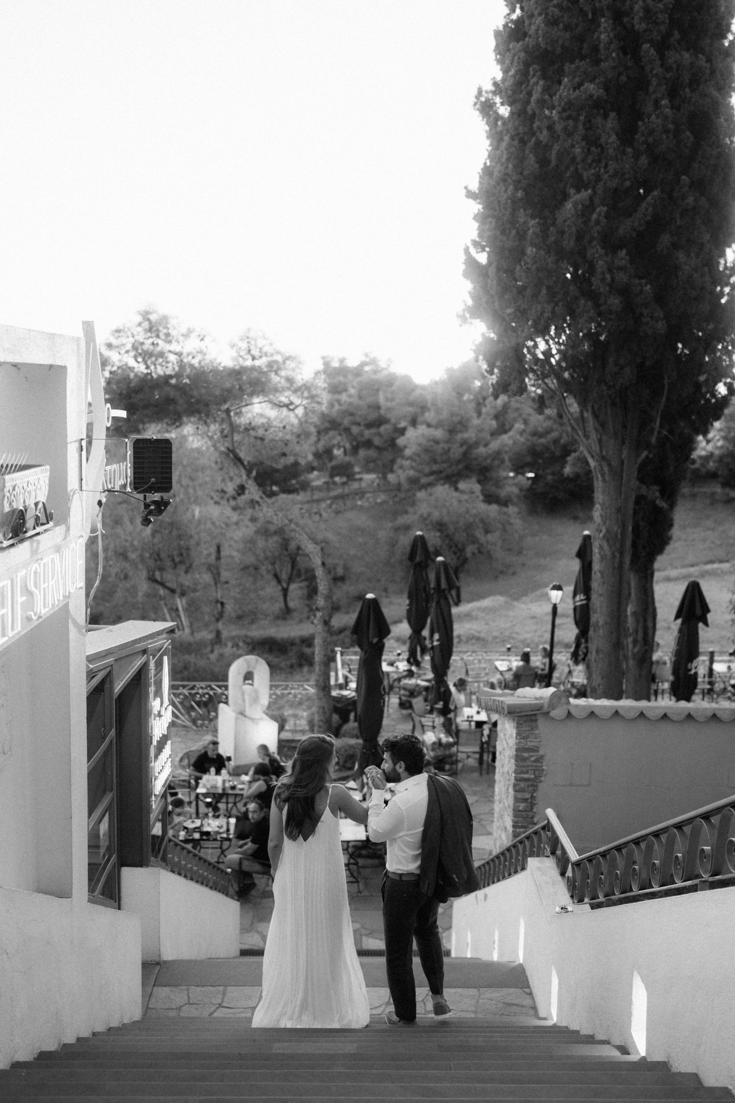 Black and white photo of a bride and groom standing on stone steps at an outdoor wedding venue surrounded by trees.