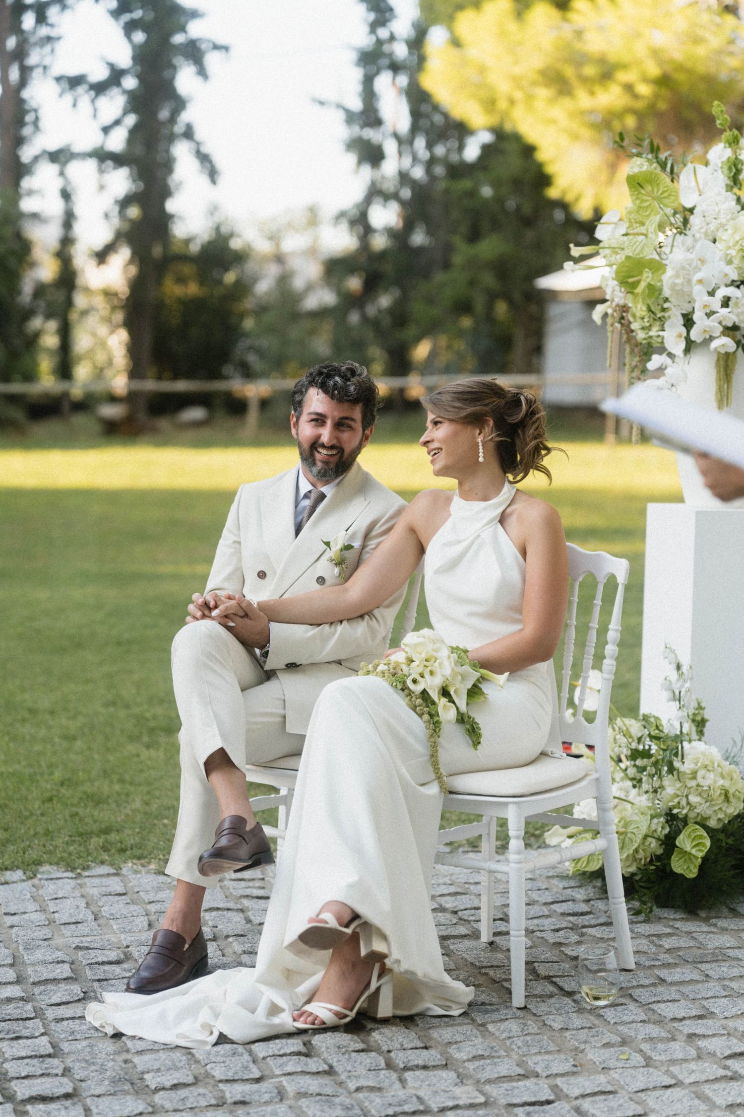 Happy bride and groom holding hands at outdoor wedding ceremony surrounded by lush greenery.