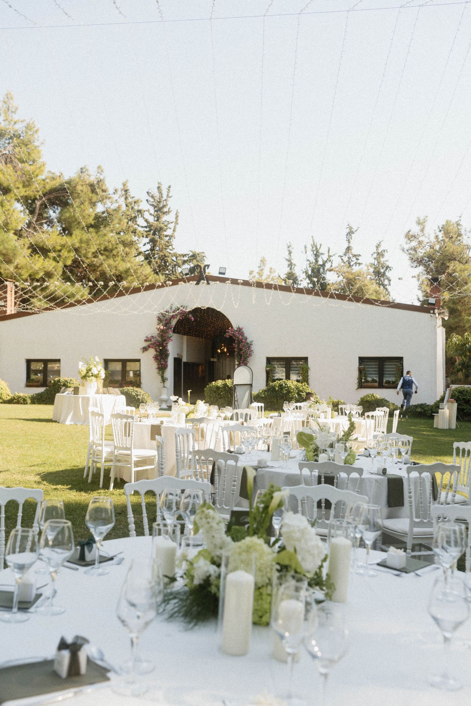 Elegant outdoor wedding reception with white tables, chairs, and floral centerpieces set on a green lawn beside a white venue.
