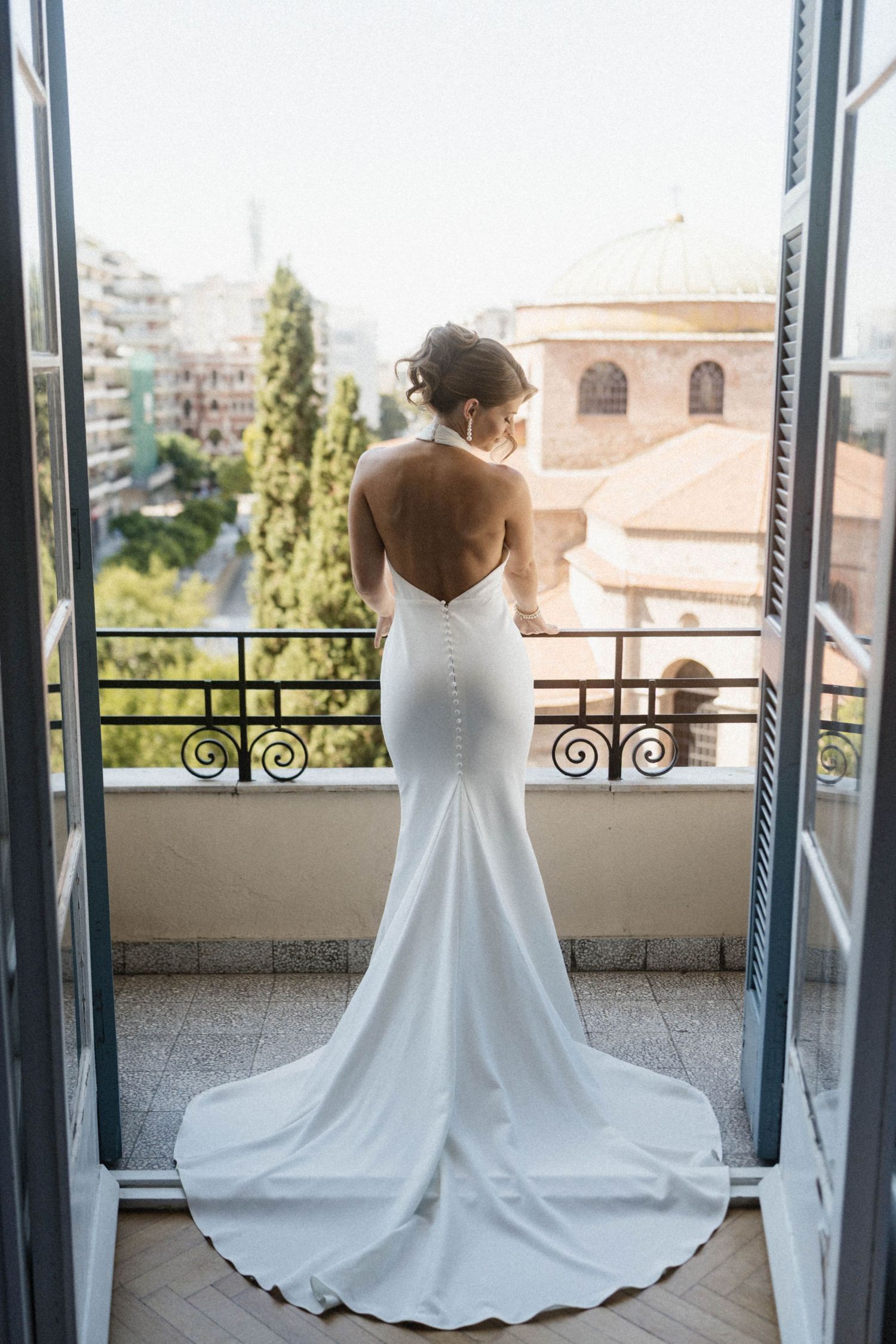 Elegant bride in backless white halter mermaid wedding gown standing on balcony overlooking historic city.