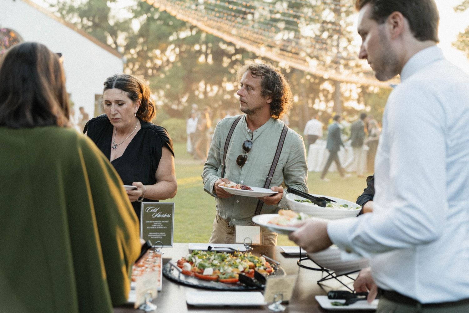 Guests serving themselves at an outdoor buffet during a garden party event with string lights in the background.