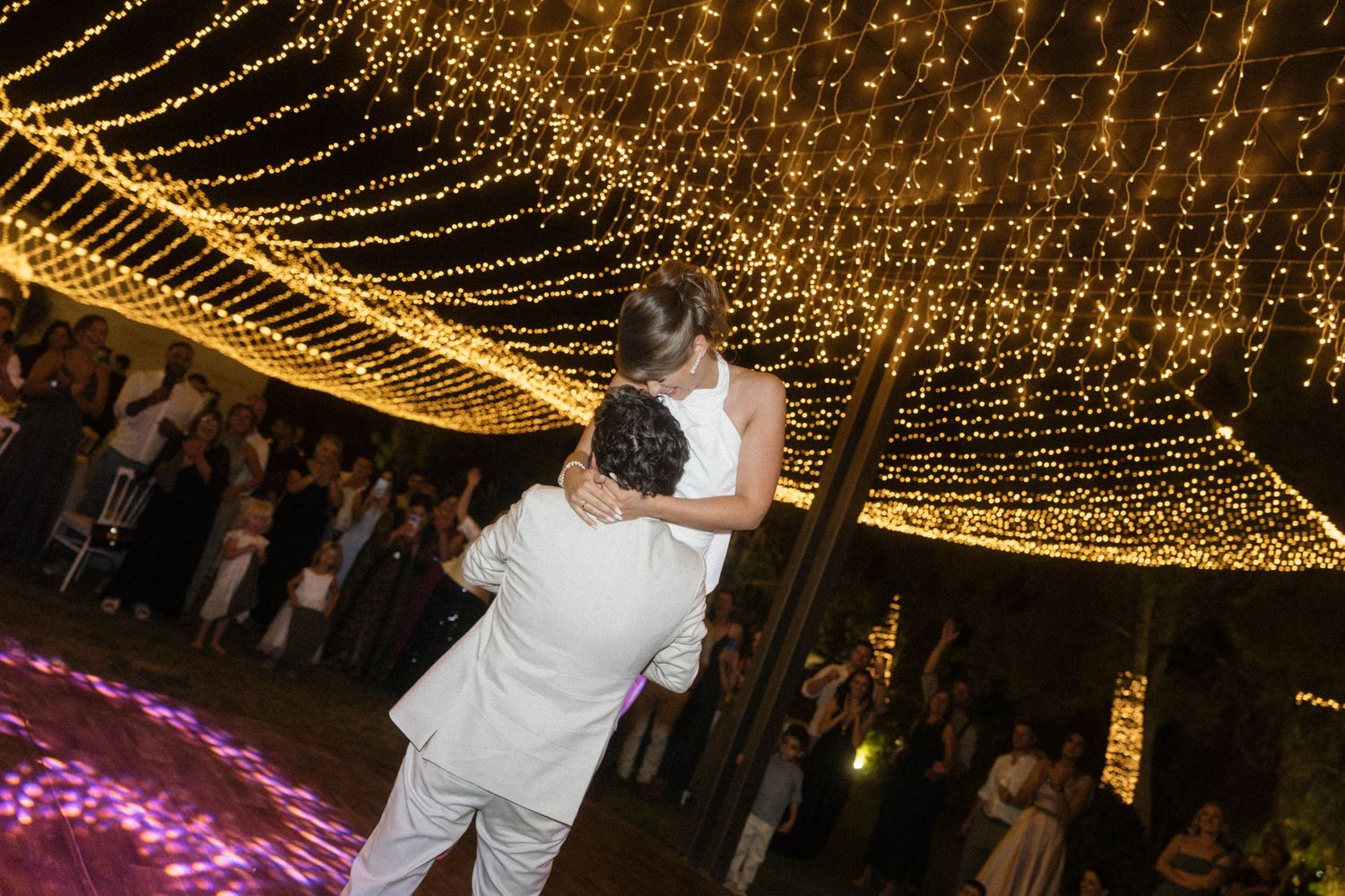 Couple shares first dance under canopy of warm fairy lights at elegant outdoor wedding reception.