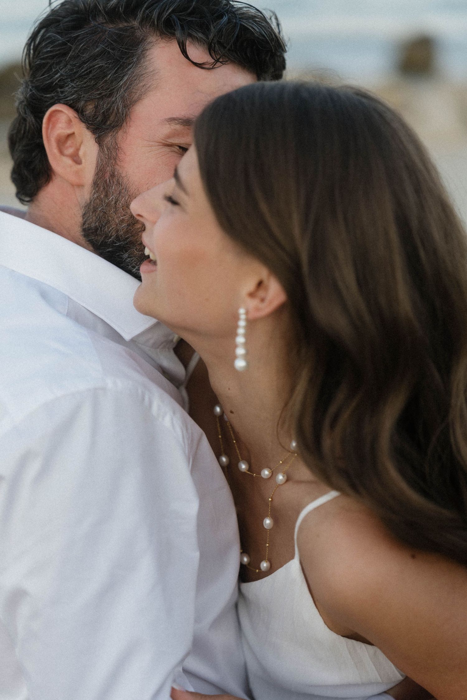 Close-up of smiling bride with pearl jewelry embracing bearded groom in white shirt at beach.