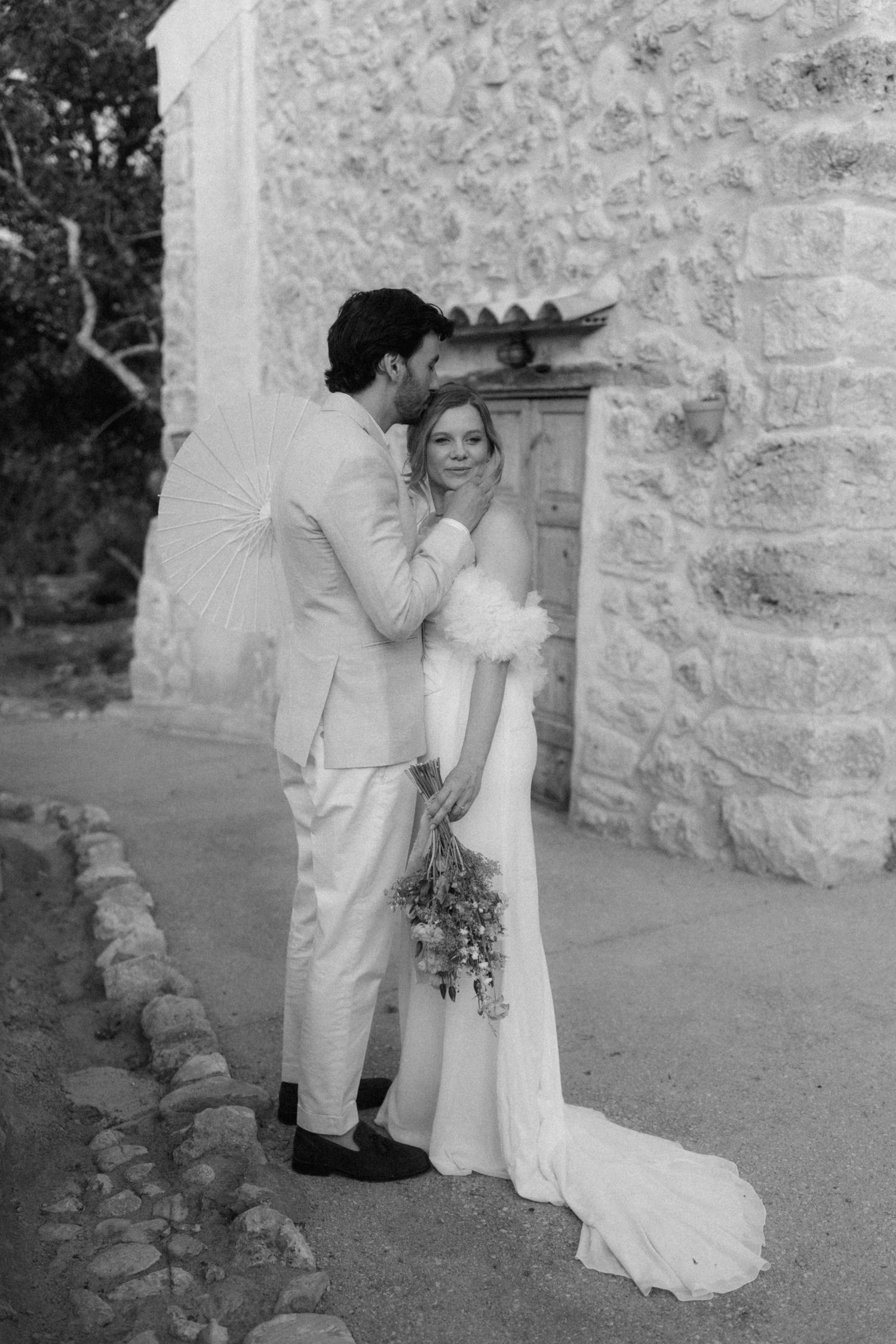 Black and white wedding photo of bride and groom embracing near stone wall, bride holding bouquet.